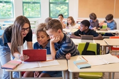 Elementary students using electronic tablet in classroom. Multi-ethnic group, girl and boys age 8-9. Teacher is explaining something to a team of two. Horizontal waist up indoors shot with copy space. This was taken in Quebec, Canada.